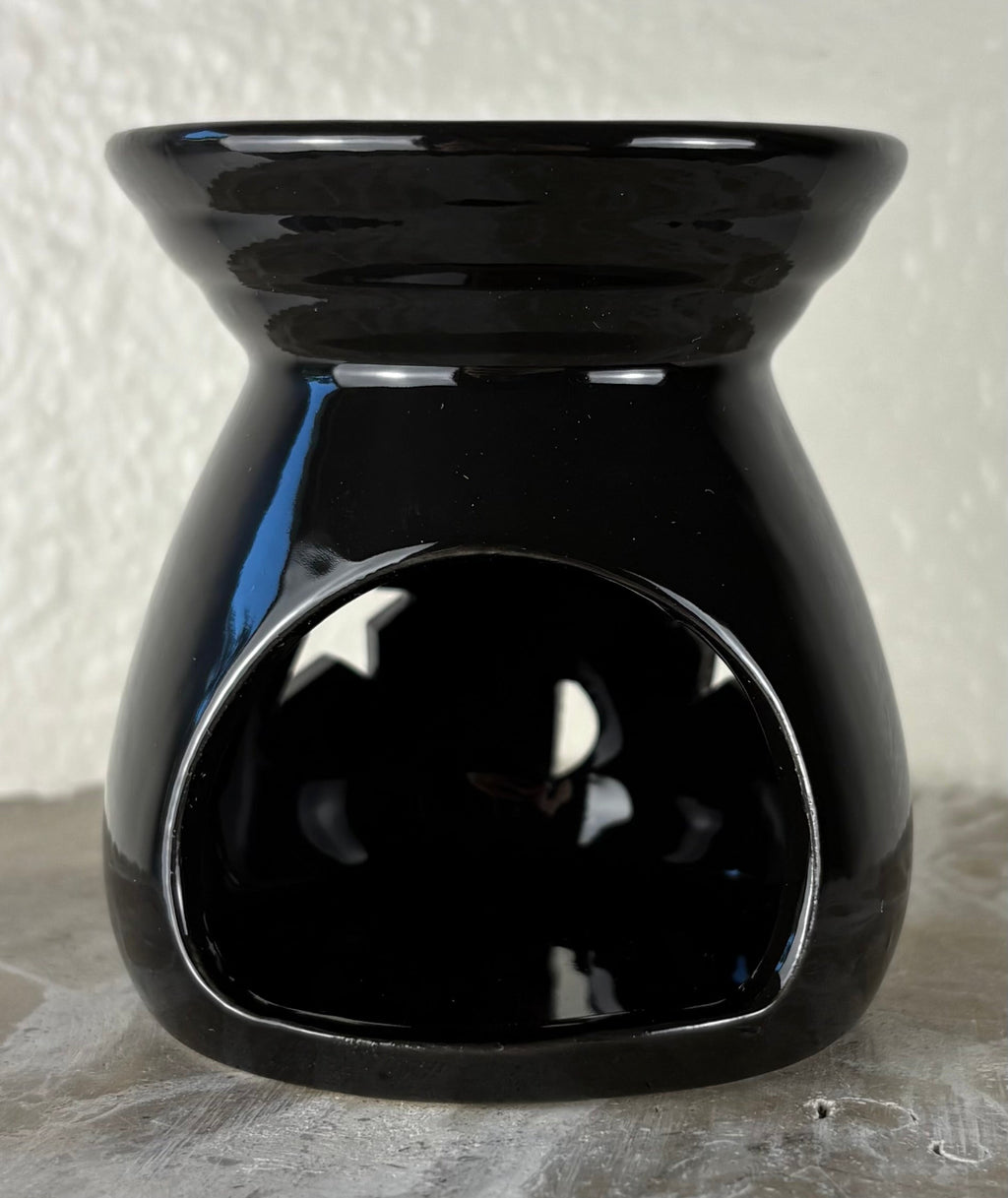 Back of a glossy black ceramic wax warmer with a wide top bowl and an open back, photographed against a light textured wall and gray surface.