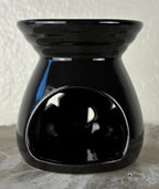 Back of a glossy black ceramic wax warmer with a wide top bowl and an open back, photographed against a light textured wall and gray surface.