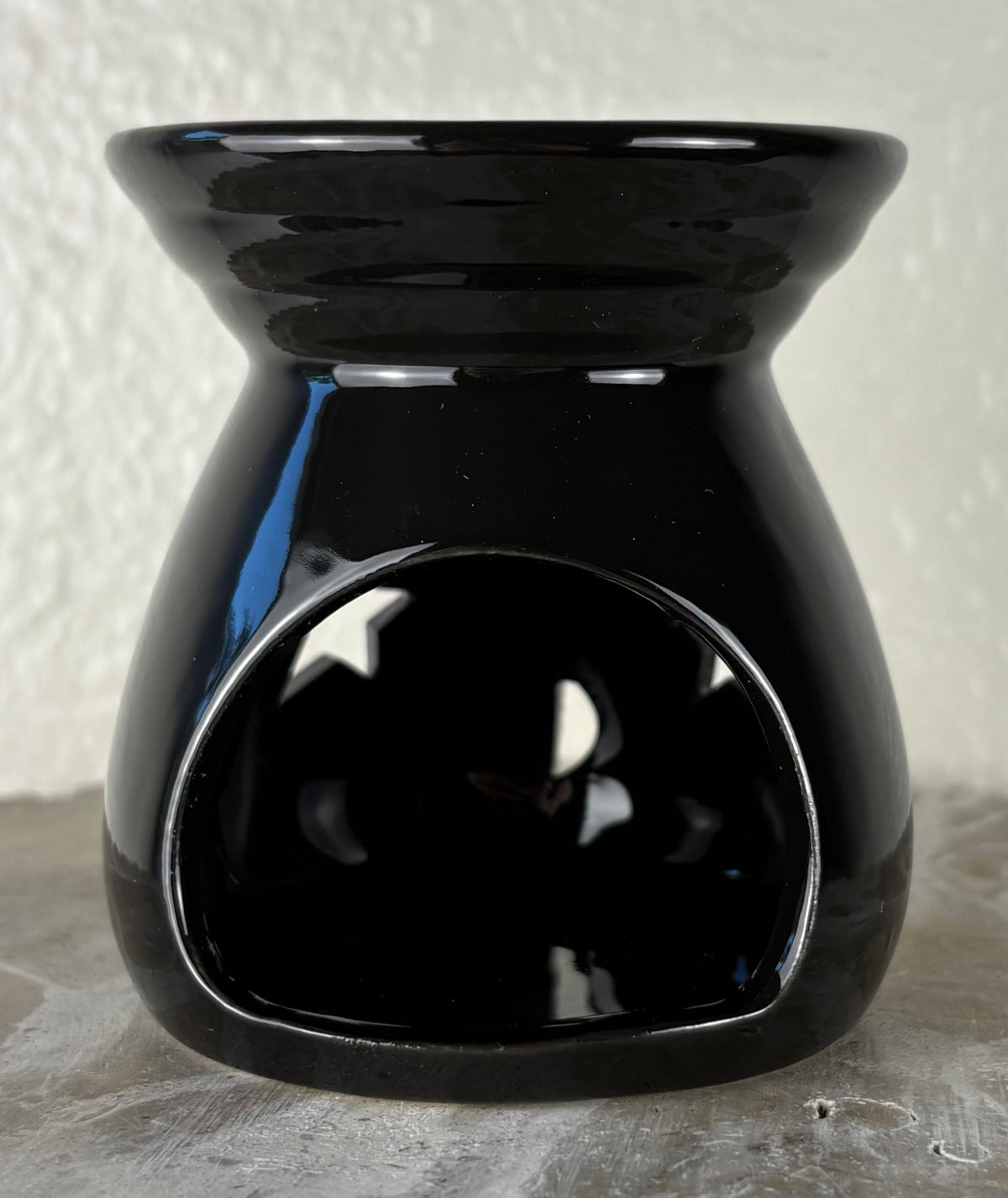 Back of a glossy black ceramic wax warmer with a wide top bowl and an open back, photographed against a light textured wall and gray surface.