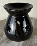 Front of a glossy black ceramic oil burner or wax warmer with star-shaped cutouts and moon-shaped cutouts, shown on a textured gray surface against a light beige wall.