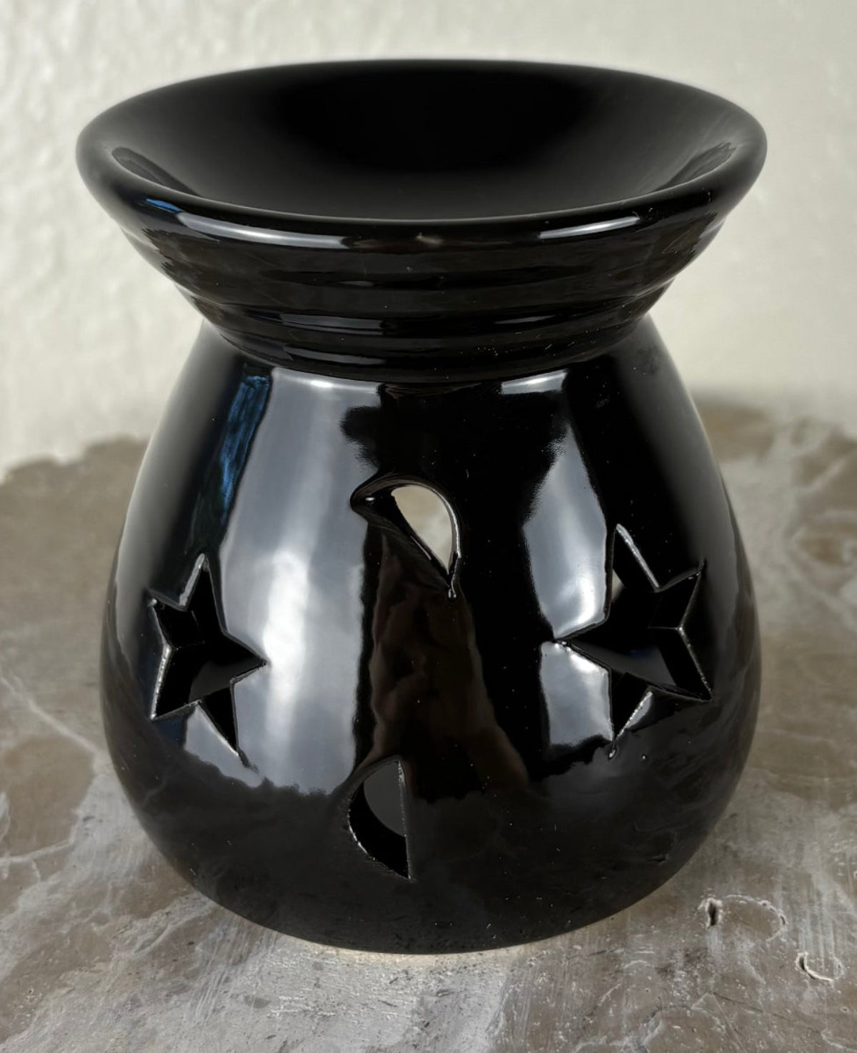 Front of a glossy black ceramic oil burner or wax warmer with star-shaped cutouts and moon-shaped cutouts, shown on a textured gray surface against a light beige wall.