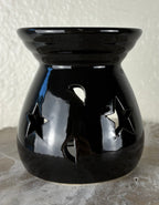 Front of a glossy black ceramic oil burner or wax warmer with star-shaped cutouts and moon-shaped cutouts, shown on a textured gray surface against a light beige wall.