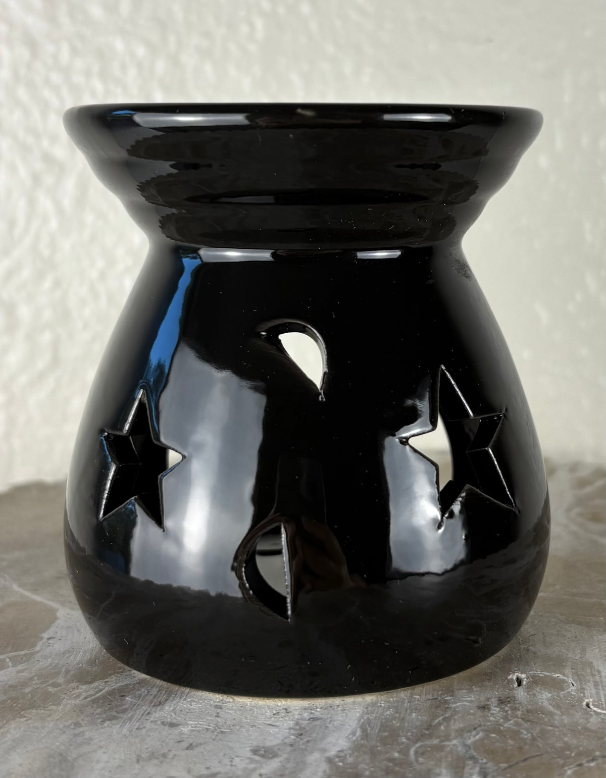 Front of a glossy black ceramic oil burner or wax warmer with star-shaped cutouts and moon-shaped cutouts, shown on a textured gray surface against a light beige wall.