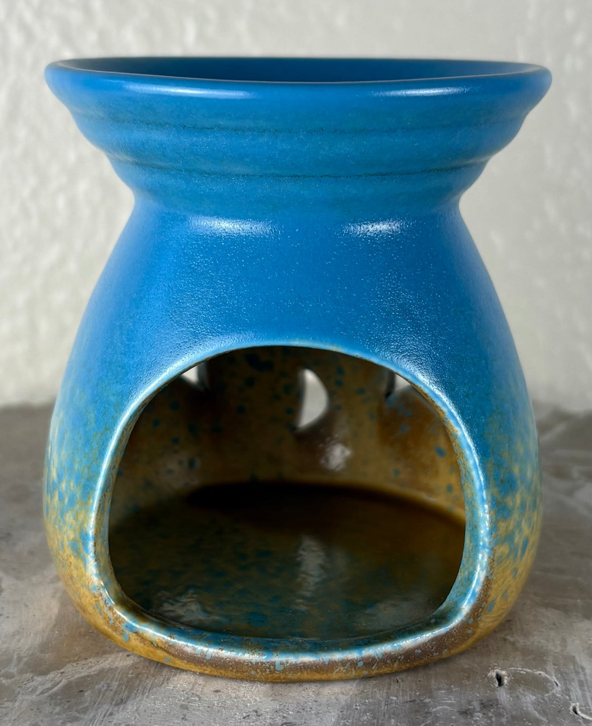 Back of a glossy blue ombre into yellow and brown ceramic wax warmer with a wide top bowl and an open back, photographed against a light textured wall and gray surface.