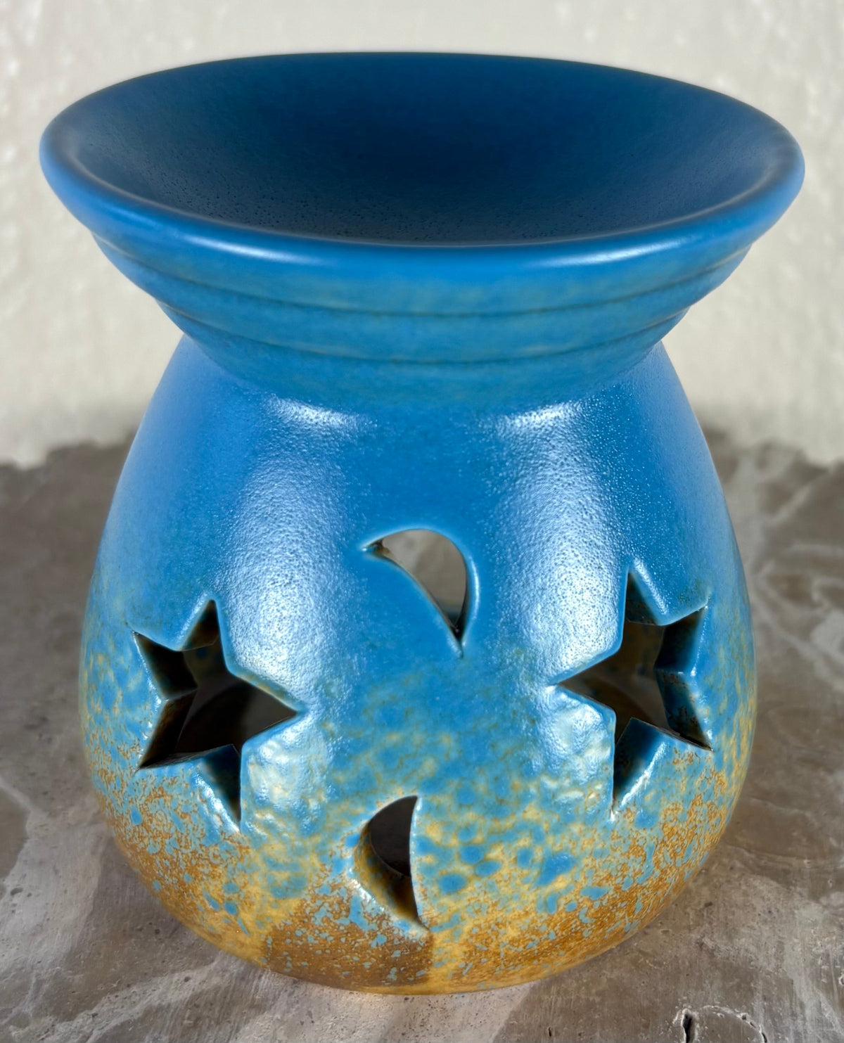 Front of a glossy blue ombre into yellow and brown ceramic oil burner or wax warmer with star-shaped cutouts and moon-shaped cutouts, shown on a textured gray surface against a light beige wall.
