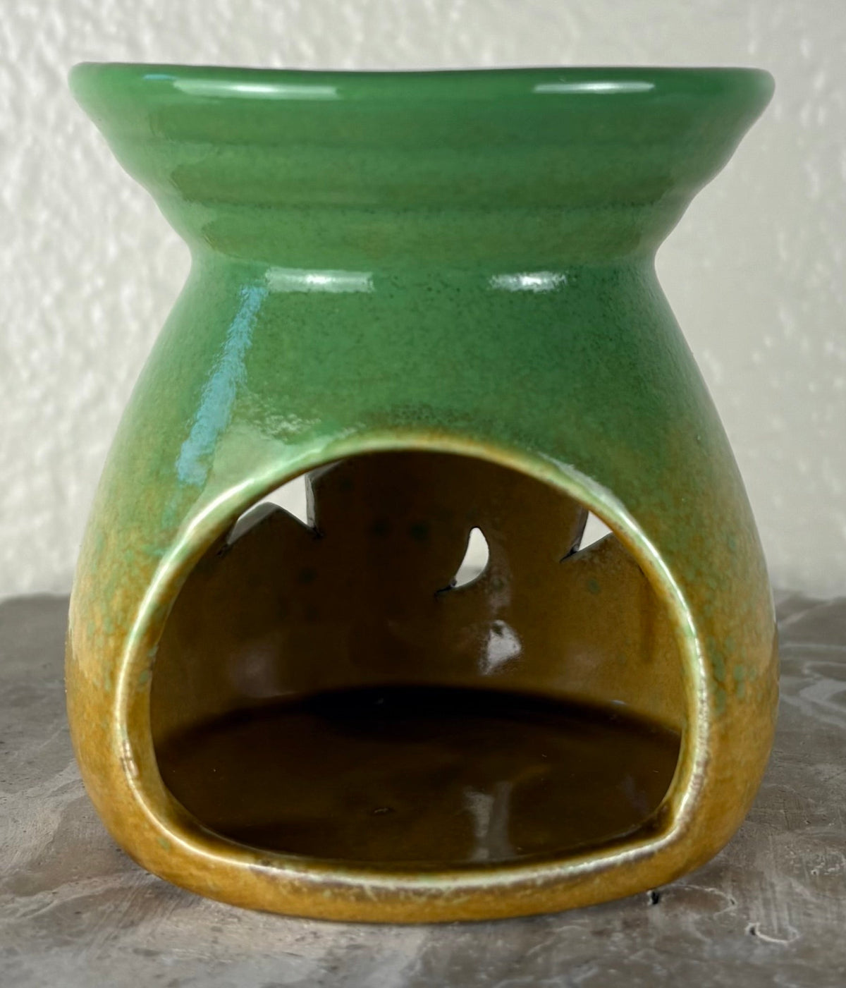 Back of a glossy green ombre into yellow and brown ceramic wax warmer with a wide top bowl and an open back, photographed against a light textured wall and gray surface.