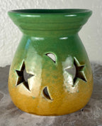 Front of a glossy green ombre into yellow and brown ceramic oil burner or wax warmer with star-shaped cutouts and moon-shaped cutouts, shown on a textured gray surface against a light beige wall.
