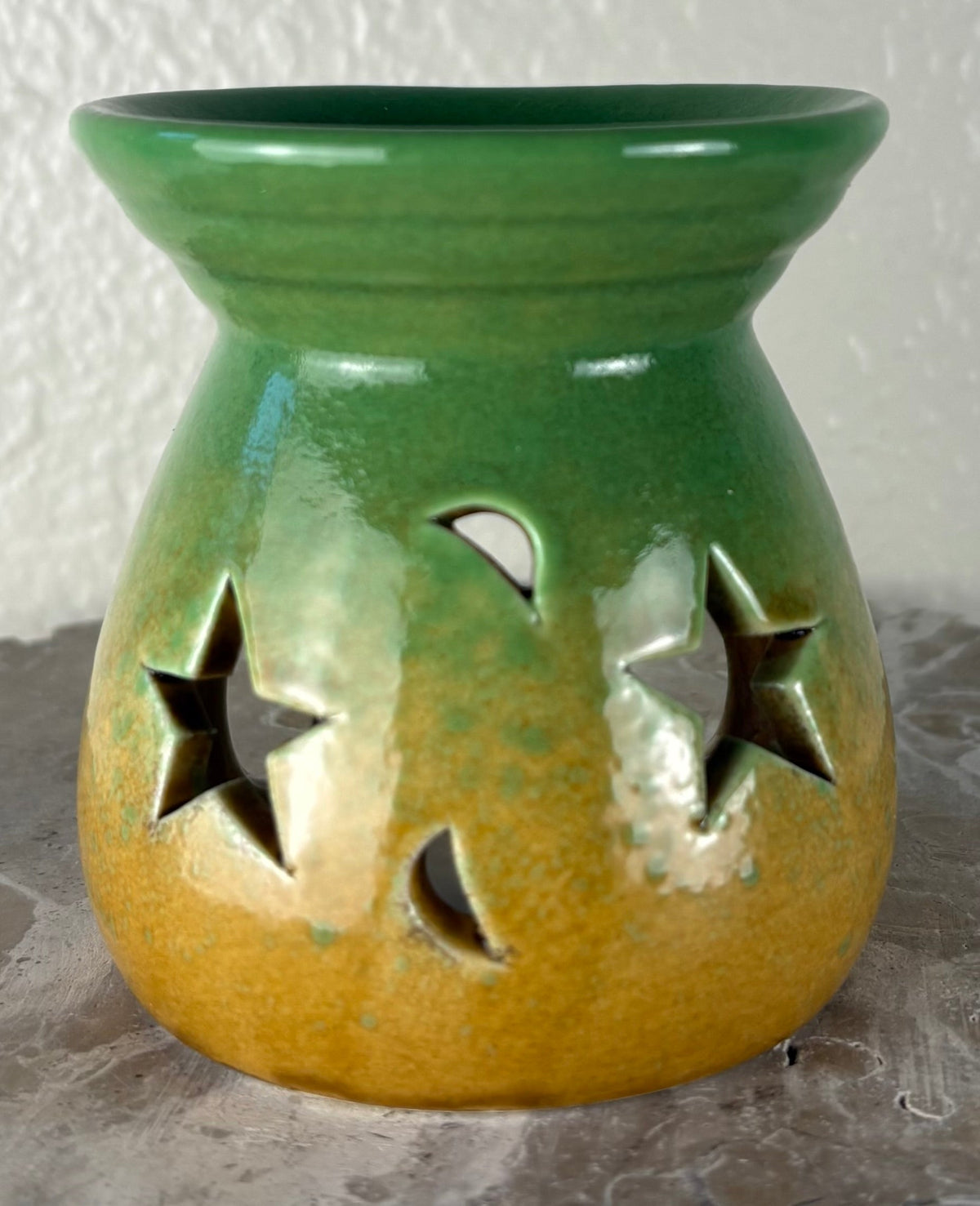 Front of a glossy green ombre into yellow and brown ceramic oil burner or wax warmer with star-shaped cutouts and moon-shaped cutouts, shown on a textured gray surface against a light beige wall.