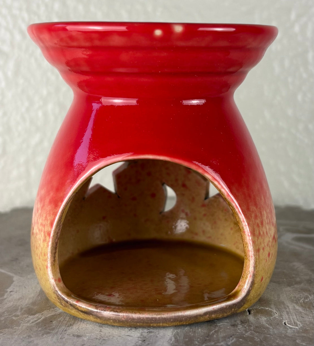 Back of a glossy red ombre into yellow and brown ceramic wax warmer with a wide top bowl and an open back, photographed against a light textured wall and gray surface.
