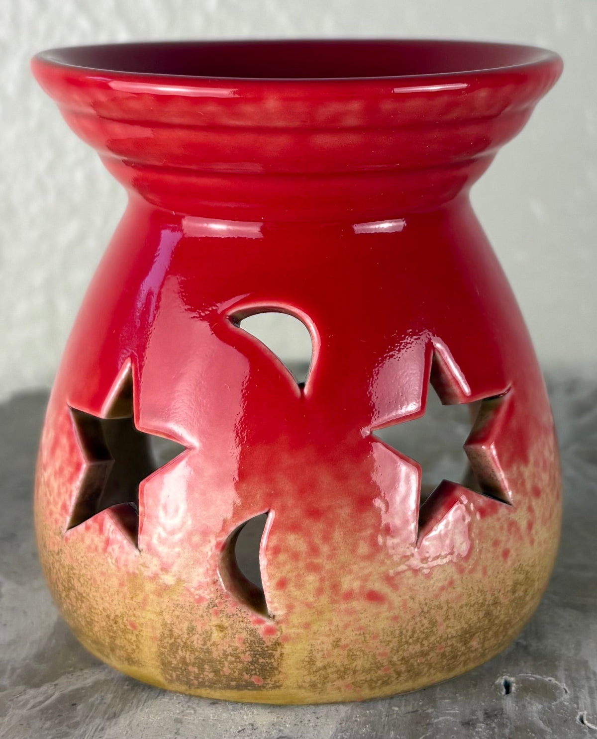 Front of a glossy red ombre into yellow and brown ceramic oil burner or wax warmer with star-shaped cutouts and moon-shaped cutouts, shown on a textured gray surface against a light beige wall.