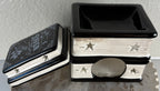 A decorative ceramic wax warmer designed to look like stacked books, with one book lid leaning against the stack. The books are black and white with star cutouts and the words 'The Raven' visible on the lid.
