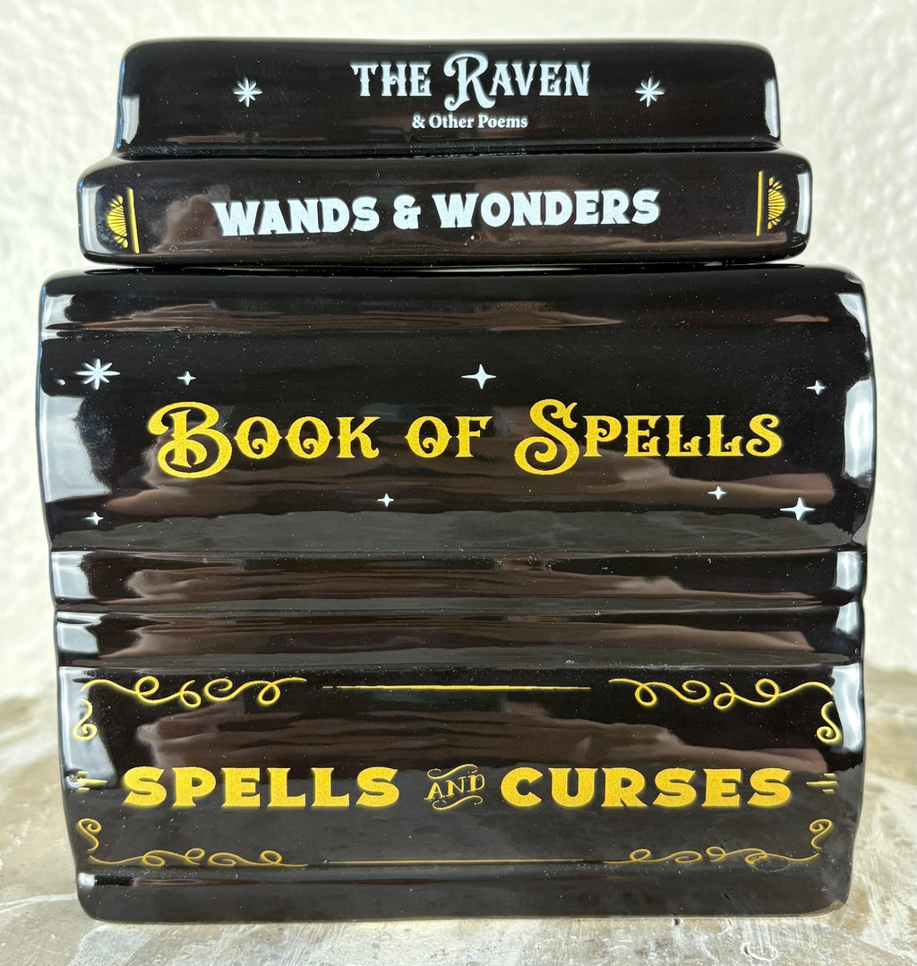 A ceramic wax warmer shaped as a stack of four glossy black decorative books or boxes with spell-themed titles: 'The Raven & Other Poems,' 'Wands & Wonders,' 'Book of Spells,' and 'Spells and Curses,' decorated with white and yellow text and star motifs. The lid is on top.