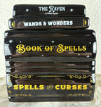 A ceramic wax warmer shaped as a stack of four glossy black decorative books or boxes with spell-themed titles: 'The Raven & Other Poems,' 'Wands & Wonders,' 'Book of Spells,' and 'Spells and Curses,' decorated with white and yellow text and star motifs. The lid is on top.