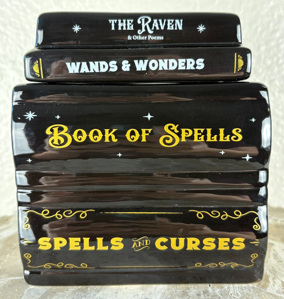 A ceramic wax warmer shaped as a stack of four glossy black decorative books or boxes with spell-themed titles: 'The Raven & Other Poems,' 'Wands & Wonders,' 'Book of Spells,' and 'Spells and Curses,' decorated with white and yellow text and star motifs. The lid is on top.