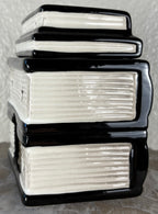 A ceramic wax warmer shaped as a stack of four glossy ceramic books with black spines and cream-colored pages, photographed close-up against a textured light background.