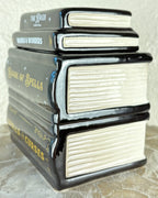 A ceramic wax warmer shaped as a stack of books featuring four stacked fantasy books: 'The Raven', 'Winds & Wonders', 'Book of Spells', and 'Curses & Curves'. The books have glossy black spines, cream-colored pages, and gold lettering, set against a neutral textured background.
