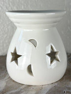 Front of a glossy white ceramic oil burner or wax warmer with star-shaped cutouts and moon-shaped cutouts, shown on a textured gray surface against a light beige wall.