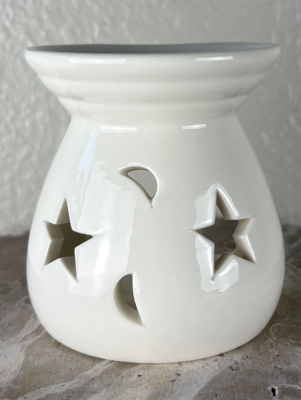 Front of a glossy white ceramic oil burner or wax warmer with star-shaped cutouts and moon-shaped cutouts, shown on a textured gray surface against a light beige wall.